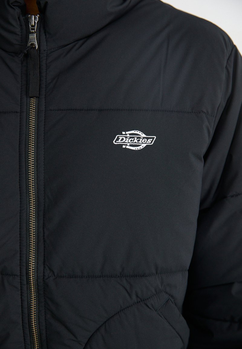 Veste matelassée noire avec un col montant, dotée d'une fermeture éclair argentée et d'un petit logo brodé blanc sur la poitrine. Texture lisse.