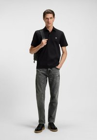 Polo nero con colletto e logo, abbinato a jeans grigi testurizzati e sneakers nere. Il modello tiene una tracolla di uno zaino.