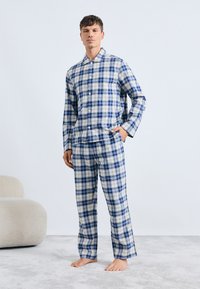 Pajamas set s karirastim vzorom v modri in beli barvi, ki vključuje srajco na gumb in ujemajoče se hlače z mehko teksturo. Model stoji bos na svetli preprogi.