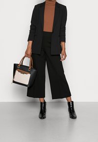 Mustapukinen bleiseri ruskean ribbitun poolopaidan päällä, yhdistettynä mustiin leveisiin housuihin ja mustiin nilkkureihin. Pitää mustaa käsilaukkua, jossa on kreeminvärinen yksityiskohta.