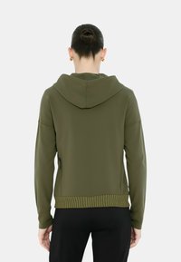 Giacca a cappuccio verde oliva con orlo a coste, tasche laterali con zip e texture liscia. Presenta una vestibilità comoda e spalle scese.