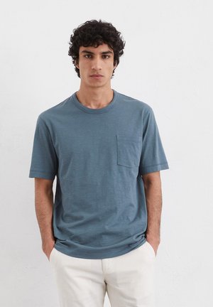 T-shirt blu a maniche corte realizzata in un tessuto morbido, con collo rotondo e taschino sul petto; indossata con pantaloni di colore chiaro.