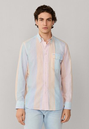 Junger Mann trägt ein pastelfarbenes, vertikal gestreiftes Hemd mit Knöpfen und hellblaue Jeans, steht frontal vor einem schlichten Hintergrund.