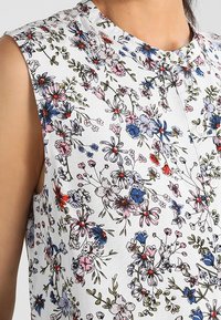 Top senza maniche floreale con sfondo bianco, caratterizzato da fiori rosa, blu e rossi; scollo rotondo e tessuto leggermente testurizzato.