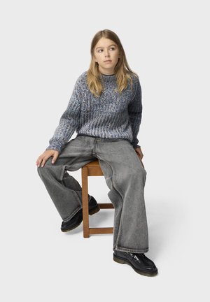 Jeune personne aux longs cheveux blonds, portant un pull en maille bleu et un jean gris large, assise sur un tabouret en bois, regardant sur le côté.