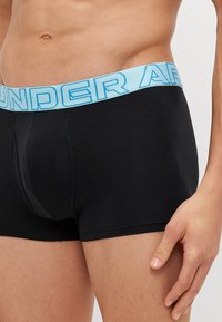 Svarte boxershorts laget av bomull, med en lyseblå elastisk midje med "UNDER ARMOUR" trykt med fete bokstaver.