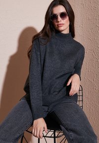 Donkergrijze trui met een hoge hals, geribbelde manchetten en tailleband, gecombineerd met bijpassende donkere jeans. Model met ronde zonnebril, zittend.