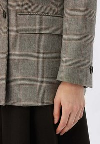 Grå rutigt kavaj med en strukturerad design, texturerat tyg och en enda svart knapp på manschetten, i kombination med en mörk kjol.