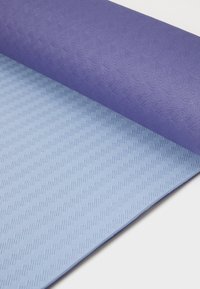 Un tapis de yoga texturé violet partiellement enroulé sur un tapis lisse bleu clair avec un motif en chevron, mettant en valeur des couleurs et des matériaux distincts.