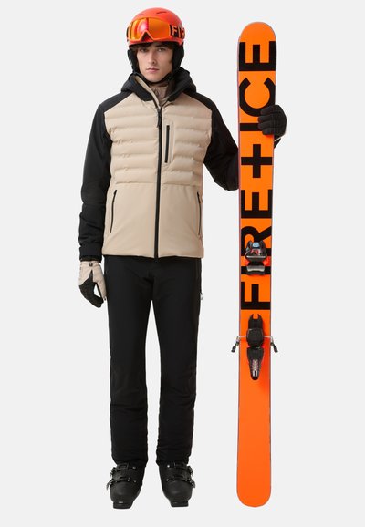 Beige und schwarze gestepptes Skijacke mit Reißverschlüssen; schwarze Skihose; hält leuchtend orangefarbene Skier mit "FIRE+ICE"-Logo; roter Helm und Skibrille.