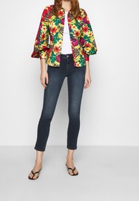 Giacca con motivo floreale e maniche a sbuffo in rosso, giallo e verde sopra una maglietta bianca, abbinata a jeans skinny scuri e infradito neri.