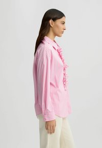 Chemise rayée rose clair avec un plastron froncé, manches longues, poignets boutonnés et coupe décontractée. Le tissu semble lisse et léger.