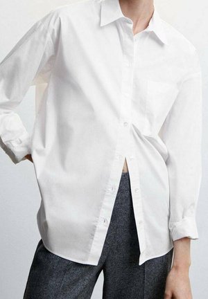 Personne portant une chemise blanche ample boutonnée avec un pantalon gris, debout devant un fond clair uni.
