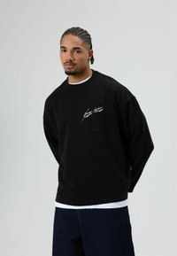 Zwarte trui met lange mouwen, een losse pasvorm, een zakdetail en witte kenmerkende tekst op de borst. Gecombineerd met donkere denim jeans.