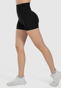 Hoge zwarte shorts gemaakt van rekbare stof; heeft twee zijzakken en een glad, gestructureerd oppervlak. Gekocht met witte sportschoenen.