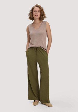 Frau mit lockigem Haar, die ein ärmelloses beige Stricktop, olivgrüne weite Hosen und gewebte beige Slippers trägt, steht mit den Händen in den Taschen.