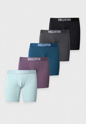 Schlanker dunkelhäutiger Mann steht barfuß mit definierten Muskeln, trägt blaue Hollister-Boxershorts vor einem schlichten weißen Hintergrund.