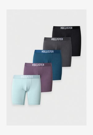 Slank mørkhudet mand står barfodet med veltrænede muskler, iført blå Hollister bokserbriefs, mod en ensfarvet hvid baggrund.