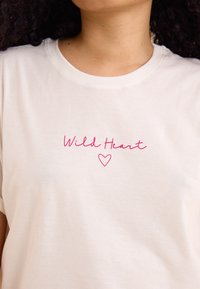 Camiseta de algodón color crema con texto en cursiva rosa "Corazón Salvaje" y un gráfico de un corazón, con cuello redondo clásico y mangas cortas.