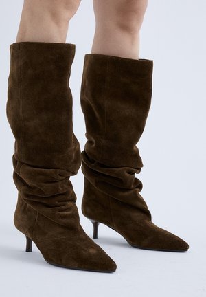 Botas marrones de ante hasta media pantorrilla, de diseño holgado, con puntera puntiaguda y tacones finos bajos, usadas sobre piernas desnudas con un fondo liso.