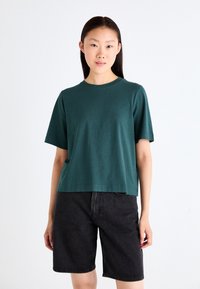 T-shirt en coton vert foncé à manches courtes, col rond et coupe décontractée. Associé à un short en denim noir. Design simple sans motifs.