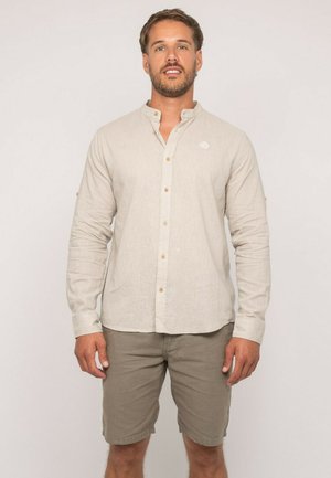 Homme debout portant une chemise beige à manches longues avec col montant et des shorts vert olive sur un fond uni.