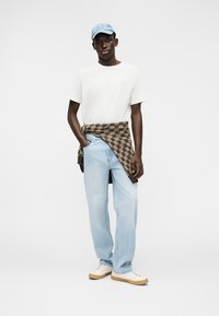 Informele outfit met een wit T-shirt, lichtblauwe spijkerbroek, een bruin geruit vest dat om de taille is gebonden, en een lichtblauwe pet.