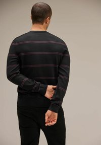 Svart stickad tröja med horisontella lila ränder, rundhalsad, med ribbad ärmslut och midja; modell vänd bort från kameran.