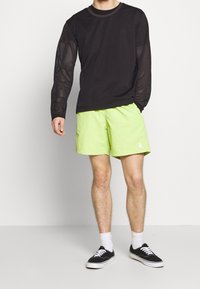 Černé síťované tričko s dlouhým rukávem a vzorovanými rukávy, doplněné světlými limetkovými šortkami. Celkový vzhled dotvářejí černé tenisky a bílé kotníkové ponožky.