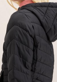 Černá prošívaná bunda s kapucí, s chevronovým prošíváním a texturovaným růžovým límcem. Materiál vypadá lehký a voděodolný.