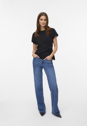 Camiseta negra de manga corta con un detalle anudado en la cintura, combinada con vaqueros de pierna ancha de color azul claro y zapatos negros de punta afilada.