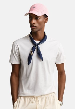 Άνδρας που φοράει λευκό μπλουζάκι, ανοιχτό μπεζ παντελόνι με κορδόνι, μπλε σκούρο φουλάρι με σχέδιο δεμένο γύρω από τον λαιμό και ροζ-άσπρο ριγωτό καπέλο.