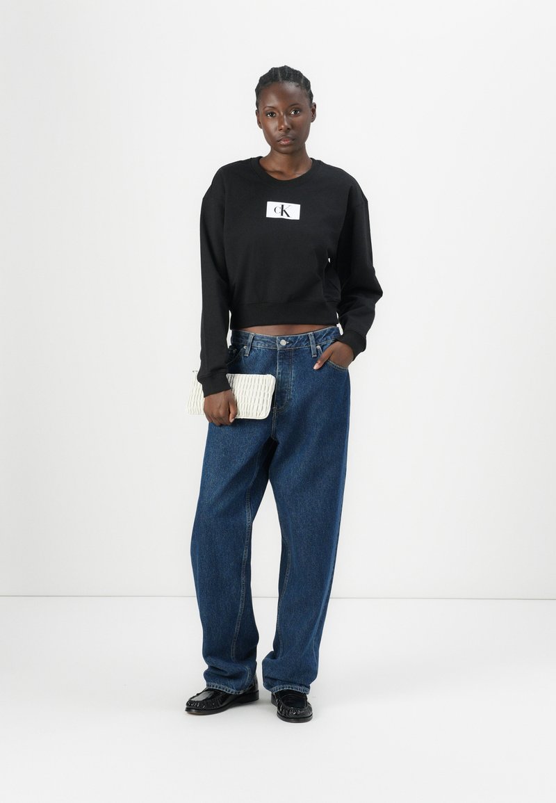 Sudadera negra corta con logo blanco, jeans de mezclilla azul sueltos y un clutch texturizado de color blanco roto. Calzado negro con un diseño detallado.