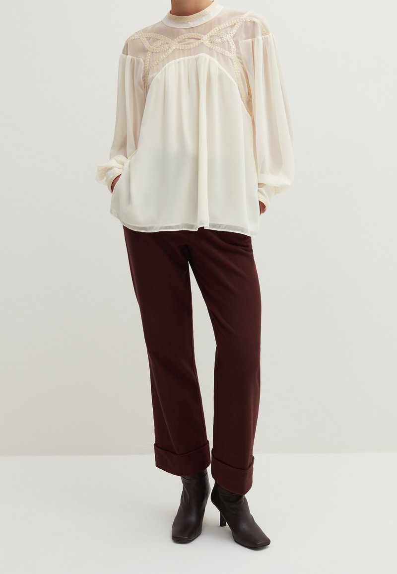 Flowy cream blouse with sheer patterned yoke and long sleeves, paired with maroon tapered pants and black heeled ankle boots.