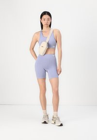 Light purple ribbed sports bra and shorts set, with a beige crossbody bag featuring a zipper and logo. Model wears beige sneakers and socks.
