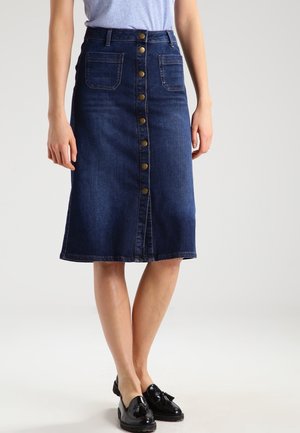 Femme portant une jupe en jean bleu foncé longueur genou avec des boutons sur le devant, des poches plaquées et des mocassins noirs à pompons.