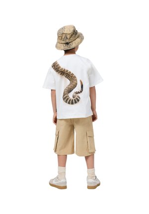 T-shirt blanche avec un graphique de serpent brun dans le dos, associé à un short cargo beige et un chapeau seau à motifs. Baskets blanches avec des accents beiges.