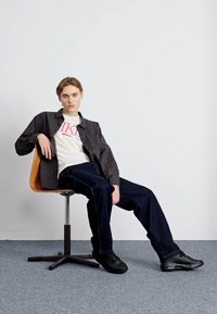 Sort og grå plaid skjorte over en hvid t-shirt med rød tekst, parret med mørkeblå brede bukser og sorte sneakers, siddende på en træstol.