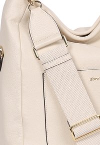 Beige Leder-Umhängetasche mit strukturiertem Oberflächen, ausgestattet mit einem breiten Stoffriemen und goldfarbenen Hardware-Accessoires. Minimalistisches Design mit Logo-Detail.