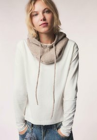 Maglietta bianca a maniche lunghe con collo alto in maglia beige, dotata di lacci. Indossata con jeans in denim blu, che mettono in risalto uno stile casual.