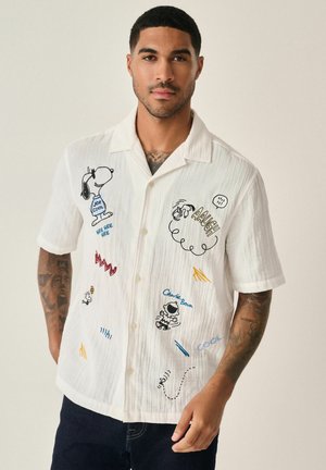Witte short-sleeve blouse met knoopsluiting, voorzien van geborduurde Snoopy- en Charlie Brown-afbeeldingen, met verschillende kleurrijke teksten en krabbels.