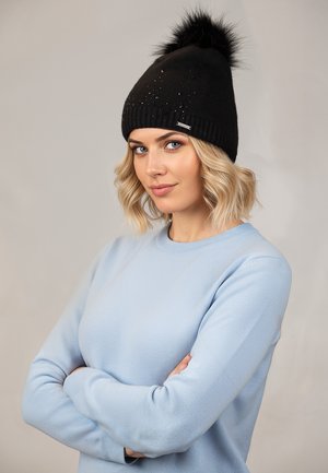 Woman with blonde wavy hair wearing a light blue sweater and black knit hat with pom-pom and small decorative studs, arms crossed, neutral background.