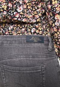 Tecido preto com padrão floral, franzido na cintura, com uns jeans de ganga cinza que apresentam um bolso costurado e uma etiqueta de marca azul.