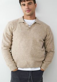 Beige Wollpullover mit Kragen, zwei Knopfverschluss, gerippten Bündchen und Saum. Trägt über einem weißen T-Shirt, kombiniert mit dunklen Hosen.