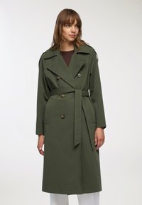 Oliwkowy płaszcz trench z podwójnym zapięciem, dużymi guzikami i wiązaniem w talii. Posiada epoletki oraz długie rękawy o dopasowanym kroju.