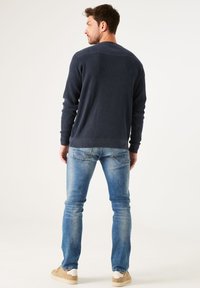 Marineblau gestrickter Pullover mit geripptem Design, Rundhalsausschnitt und kontrastierenden glatten Schulterdetails, kombiniert mit hellblauen Jeans und beigen Schuhen.