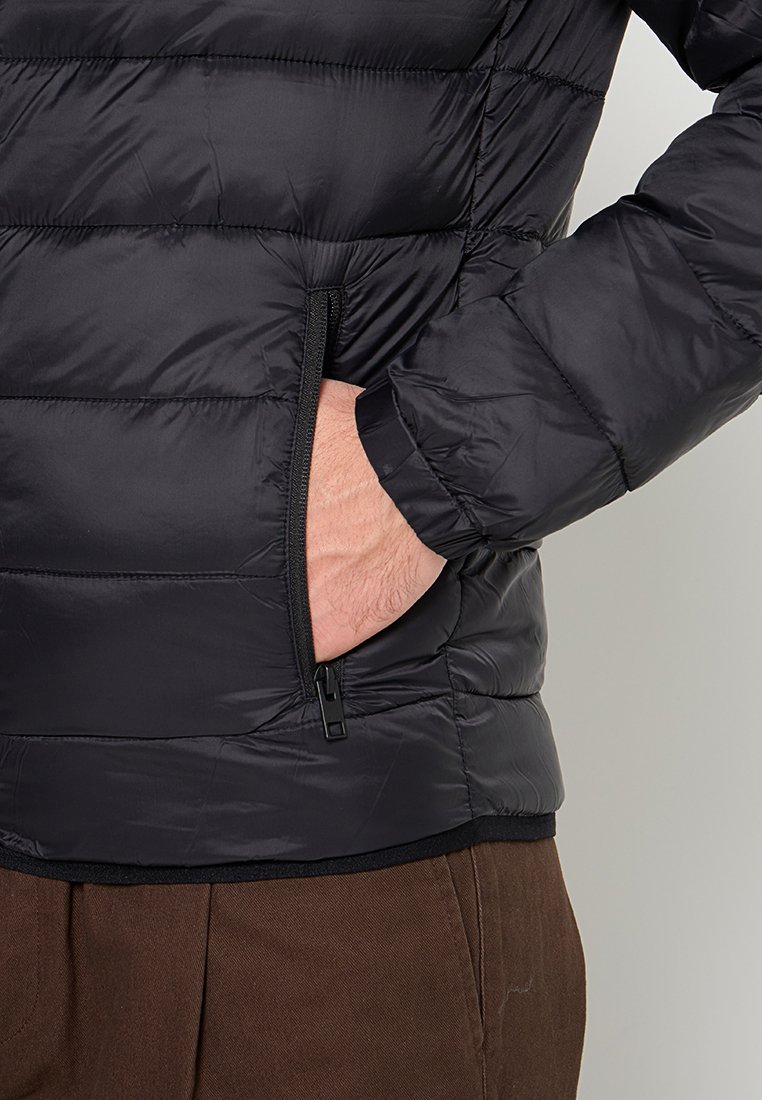 Chaqueta de plumas negra con costuras horizontales, bolsillo lateral con cremallera, textura suave y brillante, y puños elásticos. Sin marcas ni patrones visibles.