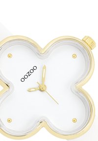 Montre dorée avec un boîtier en forme de trèfle, cadran blanc, aiguilles dorées et accents clairs. Équipée d'un bracelet blanc pour le contraste. Marquée "OOZOO."