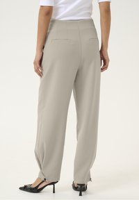 Svetle béžové kalhoty širokého střihu s hladkou texturou, vybavené dvěma zadními kapsami a knoflíkovými detaily na spodním lemu. Nosené s černými lodičkami.