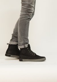 Botas de tobillo de ante negro con suela de goma gris, acento circular en el lateral y cordones grises, combinadas con jeans gris enrollados.